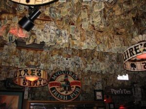 The ceiling of McGuire’s Irish Pub covered in thousands of signed dollar bills, with Irish-themed stained-glass lamps and rustic decor.