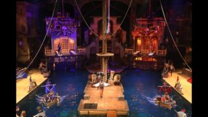 A vibrant pirate-themed dinner show featuring a large indoor lagoon, two full-sized pirate ships, and performers in costumes engaging in stunts and acrobatics.